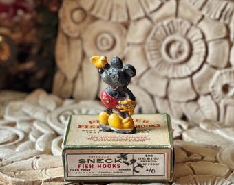 May include: A small, vintage-style Mickey Mouse figurine with black, red, and yellow paint, standing on a rectangular cardboard box labeled Pflueger Sneck Fish Hooks, 100 5621-Br, Bronzed Rust Proof, 1/0, Made in U.S.A. of American Material.