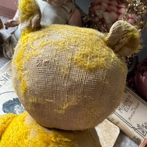 May include: Close-up of a vintage yellow teddy bear with visible wear and tear. The bear has a textured, tan body and yellow fur on its head and ears. The bear is positioned in front of a doll and a book.
