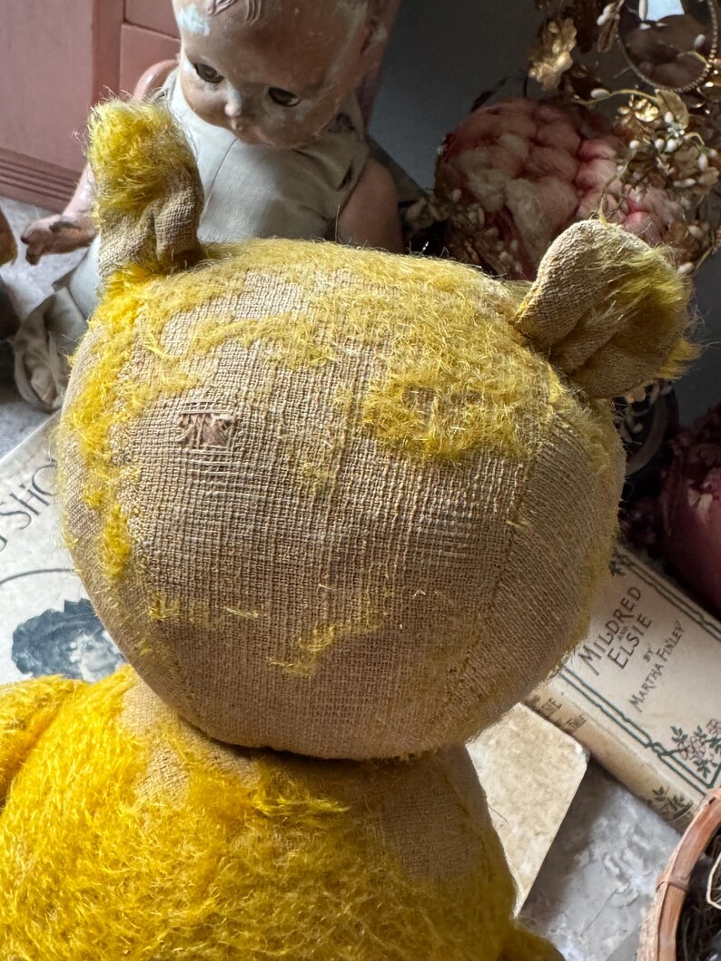 May include: Close-up of a vintage yellow teddy bear with visible wear and tear. The bear has a textured, tan body and yellow fur on its head and ears. The bear is positioned in front of a doll and a book.
