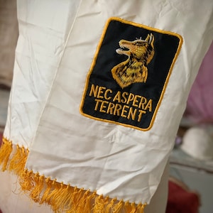 May include: White satin scarf with a gold fringe and a black embroidered patch featuring a wolf head and the text "NEC ASPERA TERRENT".