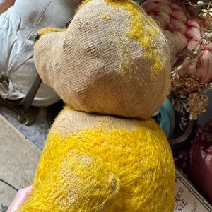 May include: A vintage teddy bear with a yellow fuzzy body and a tan head. The bear's head is made of a burlap-like material. The bear is missing an eye. The bear is a classic toy.