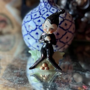 May include: A small, vintage figurine of a pixie or elf. The figurine is dressed in a black outfit with a pointed hat. It has a pale face with rosy cheeks and is standing on a green leaf base with gold accents. A blue and white patterned vase is in the background.