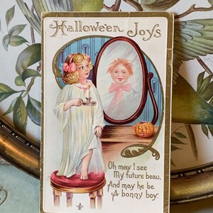 Pode incluir: Postal vintage de Halloween com uma menina de camisola segurando uma vela, olhando para um espelho. O espelho reflete um menino. O texto diz "Halloween Joys" e um poema sobre um futuro namorado.