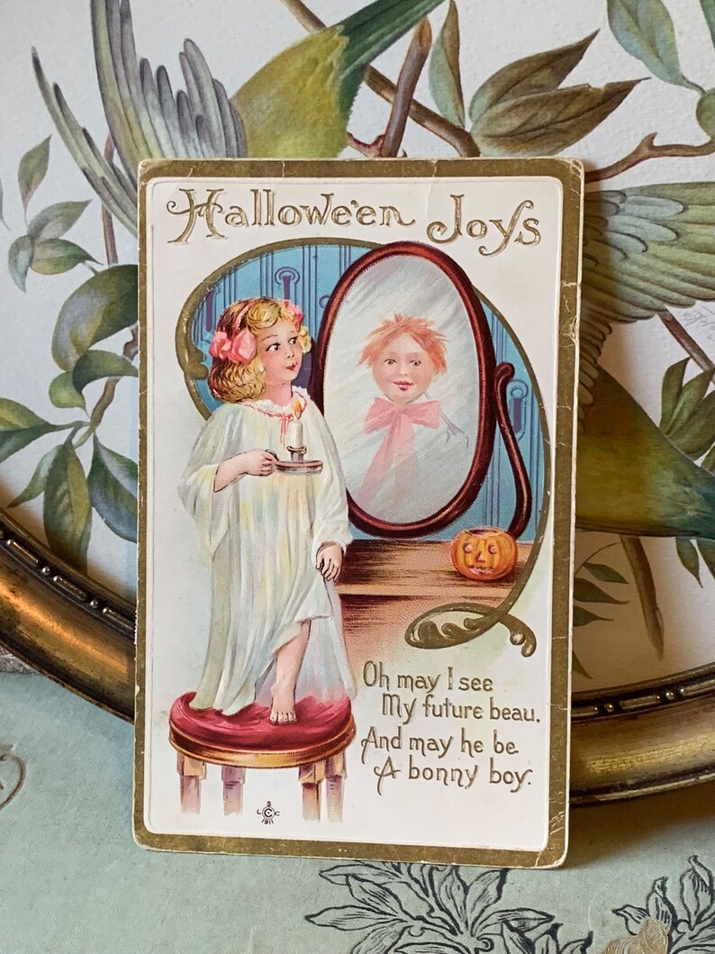 Pode incluir: Postal vintage de Halloween com uma menina de camisola segurando uma vela, olhando para um espelho. O espelho reflete um menino. O texto diz "Halloween Joys" e um poema sobre um futuro namorado.