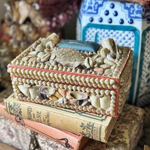 May include: A rectangular box covered in various small white, tan, and brown seashells. The lid features a central blue fabric cushion surrounded by shells. The box has a red-painted wooden frame visible between the shell layers on the sides.