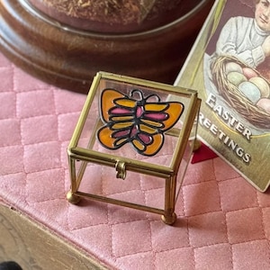 May include: A small, clear glass box with a gold trim and a hinged lid. The box contains a stained glass butterfly with orange and pink wings.