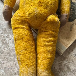May include: A vintage yellow teddy bear with brown paws and feet. The bear's fur is worn and faded, showing signs of age. The legs are positioned in a seated pose, with the feet pointing downwards. The bear is resting on a surface with aged paper.