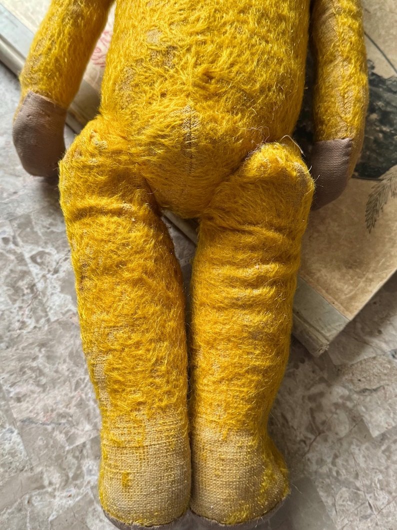 May include: A vintage yellow teddy bear with brown paws and feet. The bear's fur is worn and faded, showing signs of age. The legs are positioned in a seated pose, with the feet pointing downwards. The bear is resting on a surface with aged paper.