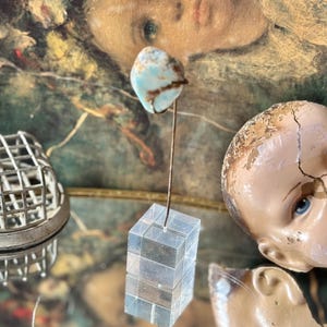 May include: A still life composition featuring a cracked doll head with blue eyes, a small turquoise and brown stone on a wire, and a clear, stacked cube. A metal floral frog is also visible.