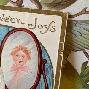 Pode incluir: Postal vintage de Halloween com o reflexo de uma crian&ccedil;a num espelho. O espelho &eacute; emoldurado em castanho escuro, e a crian&ccedil;a usa um la&ccedil;o rosa. As palavras "ween Joys" est&atilde;o em letras douradas.