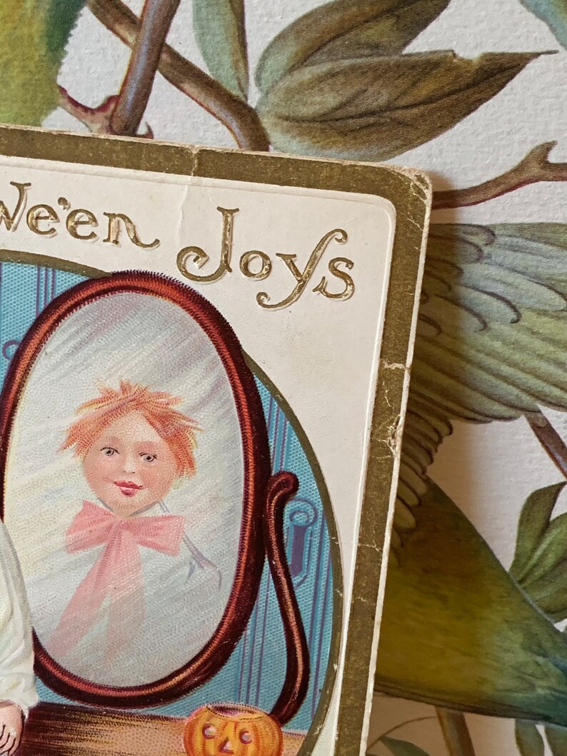Pode incluir: Postal vintage de Halloween com o reflexo de uma crian&ccedil;a num espelho. O espelho &eacute; emoldurado em castanho escuro, e a crian&ccedil;a usa um la&ccedil;o rosa. As palavras "ween Joys" est&atilde;o em letras douradas.