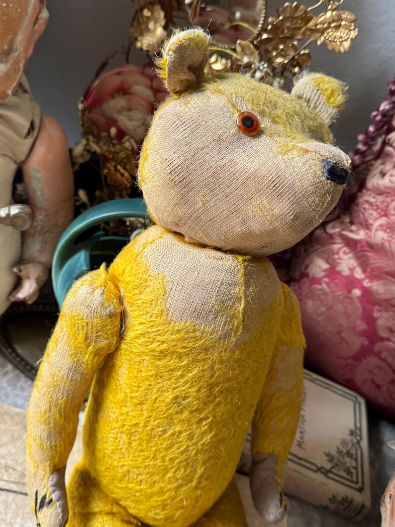 May include: A vintage teddy bear with a yellow body and a tan head. The bear has a button eye and a black nose. The bear's fur is worn, giving it a well-loved appearance. The bear is a classic toy.