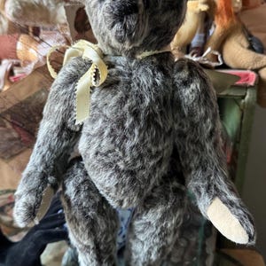 May include: A vintage-style teddy bear with gray fur and a cream-colored ribbon around its neck. The bear has a black nose and small, dark eyes. Its paws are a lighter tan color. Other antique dolls are visible in the background.