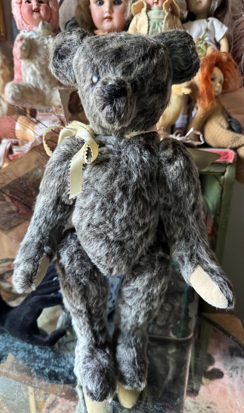 May include: A vintage-style teddy bear with gray fur and a cream-colored ribbon around its neck. The bear has a black nose and small, dark eyes. Its paws are a lighter tan color. Other antique dolls are visible in the background.