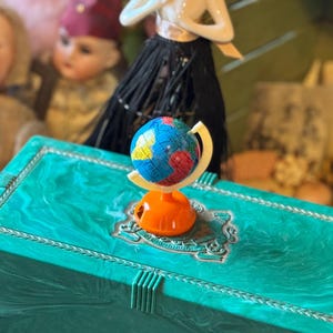 May include: A small, colorful globe on a stand sits on top of a teal green box with a decorative design. The globe is on a stand with an orange base and a white ring.