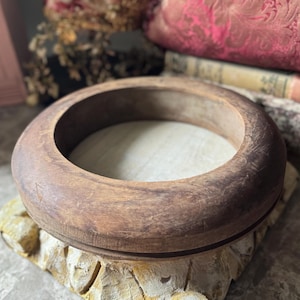 May include: A circular wooden mold with a thick, rounded rim and a smooth, light-colored interior base. The exterior of the ring features a weathered, dark brown wood grain with visible surface scratches and signs of age throughout the structure.