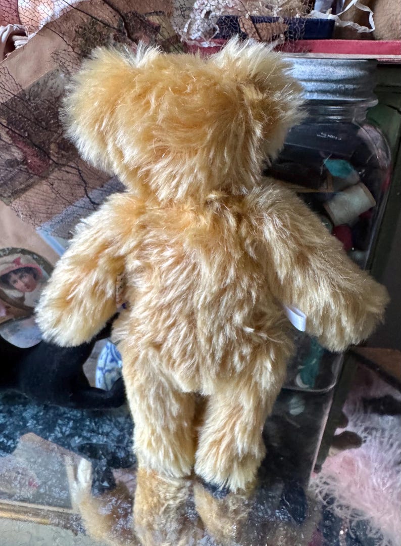 May include: A golden-colored teddy bear with a fluffy, textured appearance. The bear is viewed from the back, showcasing its arms, legs, and body. The fur is a warm, light brown color. The bear is a classic toy.