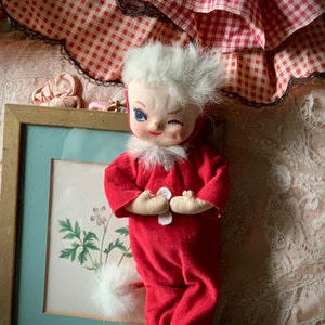 May include: A red and white plush doll with a white fur trim, winking and wearing a red jumpsuit. The doll is sitting on a white lace tablecloth with a framed picture behind it.