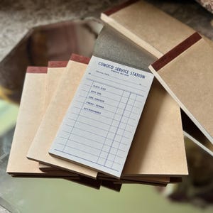 May include: A stack of rectangular paper pads with brown covers and a top-bound dark red adhesive strip. The top pad features printed blue text reading CONOCO SERVICE STATION, with lines for GAS, OIL, GREASE, TIRES, TUBES, and ACCESSORIES.