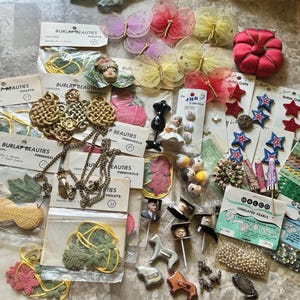 May include: A collection of assorted craft supplies including Burlap Beauties floral kits, sheer butterfly ornaments, a red fabric pincushion, star-shaped buttons, Wrights rick rack trim, Walco simulated pearls, and various small decorative figurines.