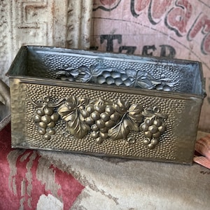 May include: Vintage rectangular metal container with embossed grape and leaf design. The container has a hammered texture and a silver-colored top. The grape design is in a gold color. The container is likely for decorative use.