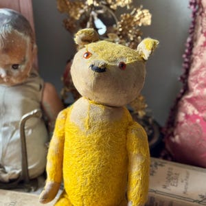 May include: A vintage yellow teddy bear with red eyes and a black nose. The bear has a tan head and ears, and its body is a bright yellow color. The bear is made of a textured fabric and has a classic, antique appearance.