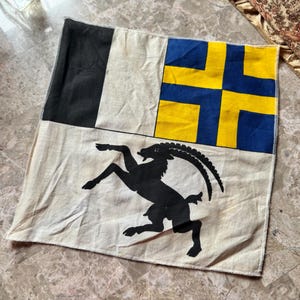 May include: A square, off-white flag with a black section, a blue and yellow cross design, and a black silhouette of a mountain goat. The flag is made of fabric and has a stitched border.