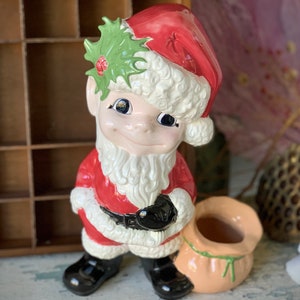 May include: A ceramic Christmas elf figurine with a red hat, white fur trim, and a green holly leaf crown. The elf is holding a black bag with a pink sack attached.