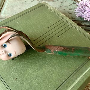 May include: A vintage green and brown garden tool with a porcelain doll head attached to the handle. The doll head has blue eyes and a painted smile.