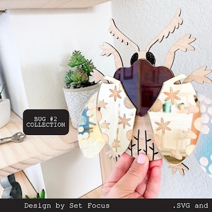 Puede incluir: Un insecto de madera con alas de espejo. Las alas están decoradas con un patrón floral y un patrón de panal. El insecto está etiquetado como "BUG #2 COLLECTION".