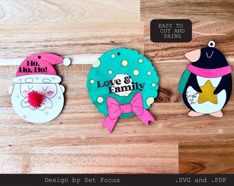 May include: Three wooden Christmas ornaments, ready to be painted. The ornaments feature a Santa Claus face, a wreath with the text "Love & Family", and a penguin holding a star. The text "Easy to cut and paint" is visible in the top right corner.