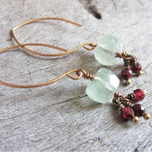 May include: A pair of dangle earrings with a light green gemstone and red gemstone accents. The earrings are made with copper wire and have a simple, elegant design.