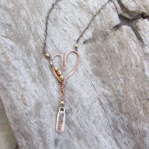 May include: A copper-colored heart pendant necklace with a delicate chain. The heart is formed from wire and adorned with small gold-colored beads. A small tag with the word "Believe" hangs below the heart. The necklace rests on a wooden surface.