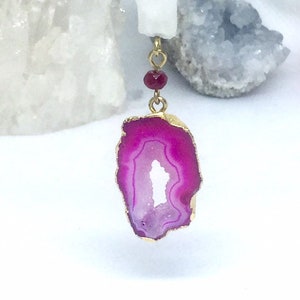 May include: A pendant necklace featuring a vibrant, fuchsia-colored agate slice with a gold-plated edge. The pendant is suspended from a delicate gold chain with small, red gemstone accents. The background includes white and gray crystal formations.