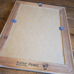 May include: The back of a wooden picture frame with a brown cardboard backing. The frame has a white label with the text "Kater Pawel recycled products" and a small black and white illustration of a cat.