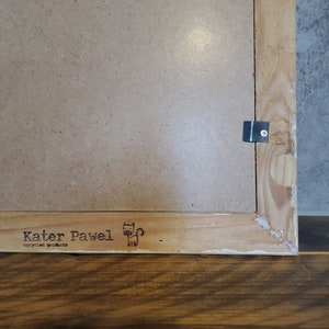 May include: A close-up of the back of a wooden picture frame with a metal hanging hook. The frame has a brown paper backing with the text "Kater Pawel recycled products" and a small cat illustration.