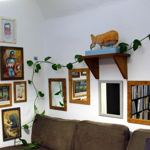May include: A white wall with a variety of framed artwork and mirrors. The artwork includes a painting of a man in a superhero costume, a painting of a forest scene, and a painting of a whale. There is also a plant hanging from the ceiling.