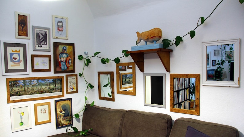 May include: A white wall with a variety of framed artwork and mirrors. The artwork includes a painting of a man in a superhero costume, a painting of a forest scene, and a painting of a whale. There is also a plant hanging from the ceiling.