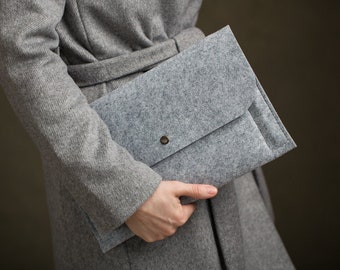 May include: A rectangular light gray felt document sleeve or clutch bag with a fold-over flap and a single centered metallic snap button closure. The material has a visible heathered texture and the design features clean, straight stitched edges.