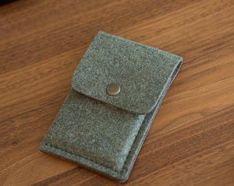 May include: A rectangular olive green felt pouch with a fold-over flap secured by a single metallic snap button. The pouch features visible stitching along the edges and a textured, soft-touch fabric construction, designed for holding small items.
