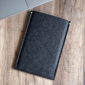 Può includere: Una custodia per laptop in feltro grigio scuro con dettagli verdi sugli angoli. La custodia rettangolare è su una superficie di legno, accanto a un laptop.