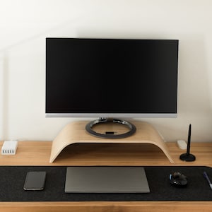Peut inclure: Un écran d'ordinateur sur un support en bois clair incurvé, posé sur un bureau en bois avec un sous-main en feutre gris foncé. L'ensemble comprend un smartphone, un ordinateur portable fermé, une souris, un stylet, un stylo et une unité centrale.