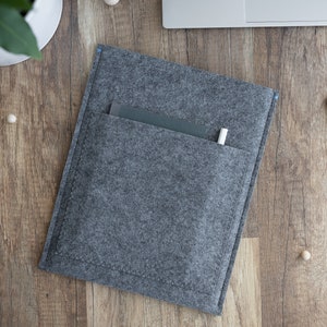 May include: A rectangular dark grey felt sleeve featuring a large front pocket with visible stitching. A dark grey notebook and a white pen are tucked inside the pocket. The sleeve has a simple design with blue thread accents at the top corners.