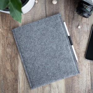 May include: A rectangular grey felt sleeve for a tablet or laptop, featuring a stitched edge and a slim profile. A white and black stylus pen rests along the right side of the sleeve, which is positioned on a wooden surface near a smartphone.