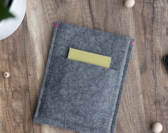 May include: A gray felt phone sleeve with a front pocket, holding a yellow card. The sleeve has pink stitching along the edge. The background is a wooden surface with a plant and a smartphone.