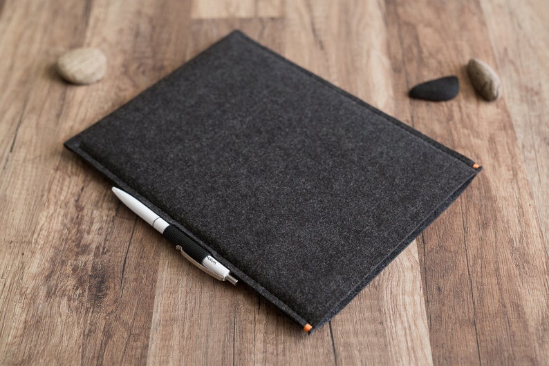 Peut inclure: Une pochette rectangulaire en feutre gris fonc&eacute; avec des bords cousus et de petits accents orange. Un stylo &agrave; bille blanc et noir est ins&eacute;r&eacute; dans la poche avant de la pochette. Le mat&eacute;riau en feutre a un aspect textur&eacute; et est plat.