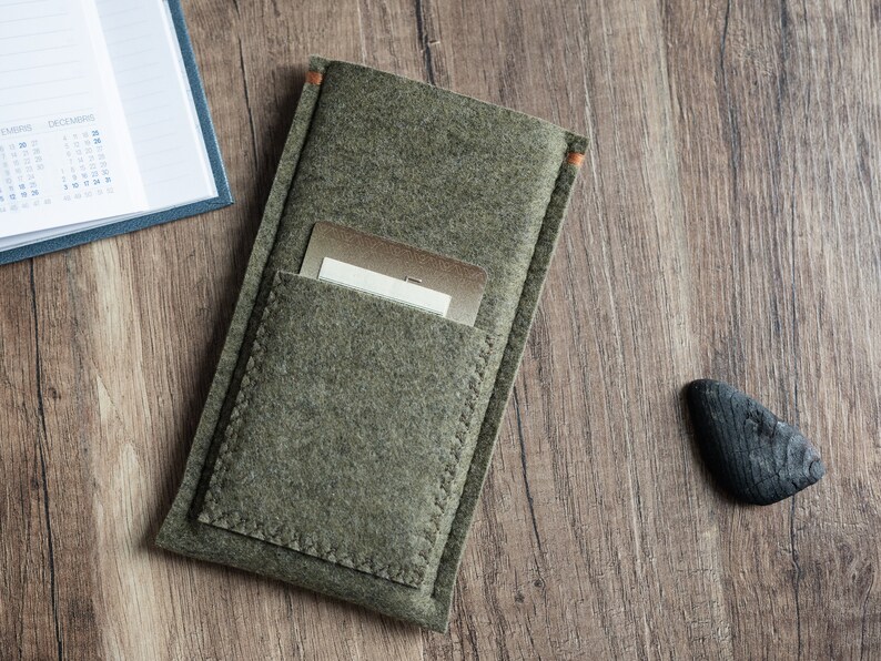 May include: An olive green felt sleeve case featuring a rectangular front pocket with visible stitching along the edges. A metallic gold card is partially inserted into the pocket. The sleeve has small orange thread accents at the top corners.