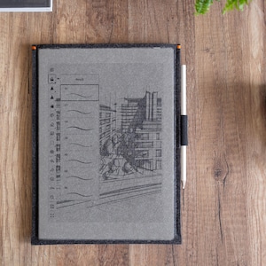 Peut inclure: Une tablette num&eacute;rique grise dans une housse en feutre fonc&eacute;, affichant un croquis architectural dessin&eacute; &agrave; la main et une barre d'outils verticale. Un stylet blanc est maintenu par une boucle &eacute;lastique noire sur le c&ocirc;t&eacute; de la housse en feutre.
