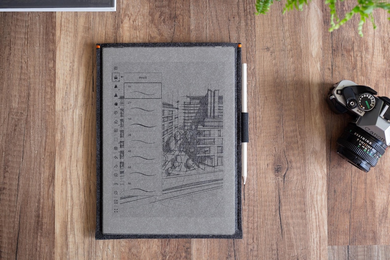 Peut inclure: Une tablette num&eacute;rique grise dans une housse en feutre fonc&eacute;, affichant un croquis architectural dessin&eacute; &agrave; la main et une barre d'outils verticale. Un stylet blanc est maintenu par une boucle &eacute;lastique noire sur le c&ocirc;t&eacute; de la housse en feutre.