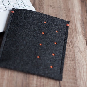 May include: A gray felt laptop sleeve with orange stitching and decorative stitching. The sleeve is lying on a wooden surface.